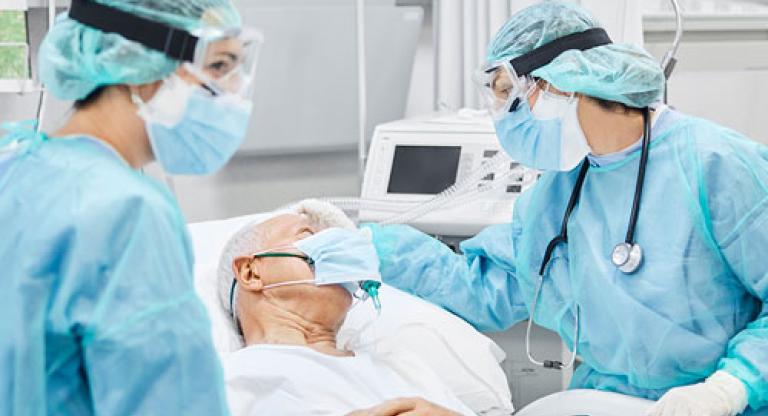 Two health care providers, wearing blue scrubs stand with a patient in the ICU 