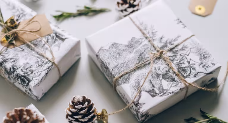 Holiday gifts wrapped in white paper and twine, surrounded by pinecones and greenery.