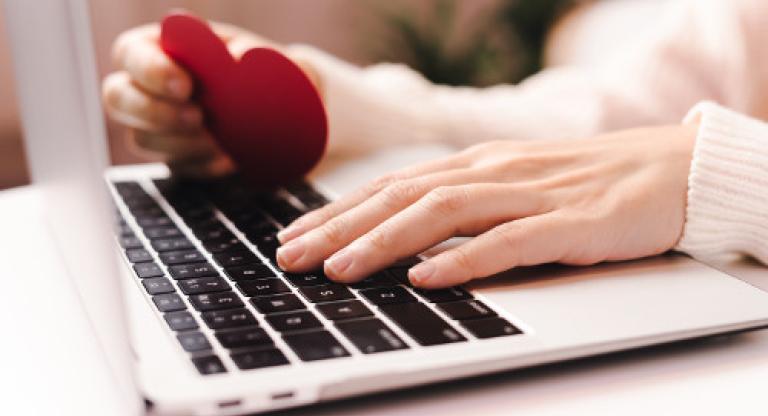 hands resting on a laptop keyboard, one is holding a red paper heart.