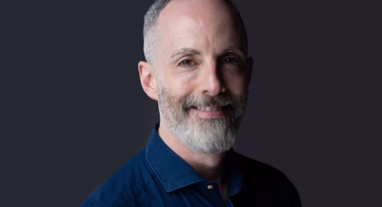 Medium close image of a smiling man with short greying hair and beard, wearing a dark blue shirt, standing against a grey background.