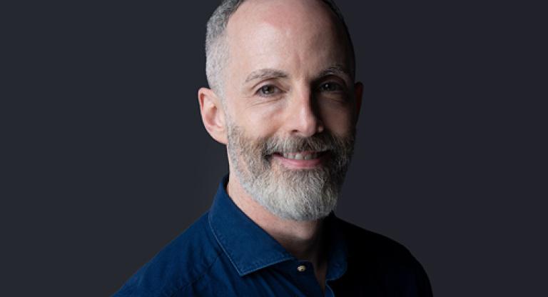Medium close image of a smiling man with short greying hair and beard, wearing a dark blue shirt, standing against a grey background.