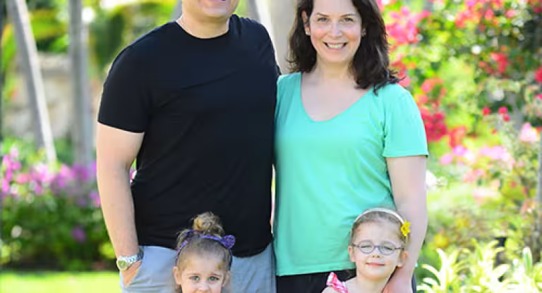 Medium long shot of a smiling family of four standing in a garden. Father wears a black shirt, mother wears a green shirt. Two young girls stand in front of them wearing pink. 