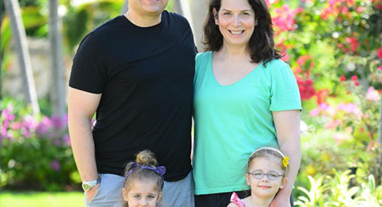 Medium long shot of a smiling family of four standing in a garden. Father wears a black shirt, mother wears a green shirt. Two young girls stand in front of them wearing pink. 