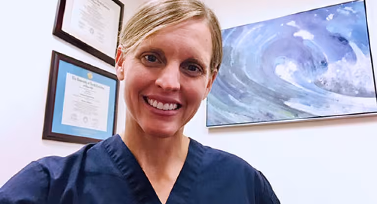 Medium close shot of a smiling blond woman, with her hair pulled back and wearing blue scrubs, sitting in an office with art and framed diplomas in the background.