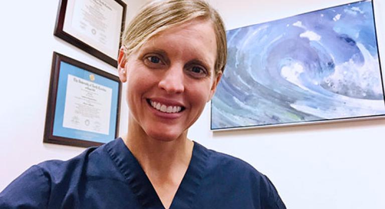 Medium close shot of a smiling blond woman, with her hair pulled back and wearing blue scrubs, sitting in an office with art and framed diplomas in the background.