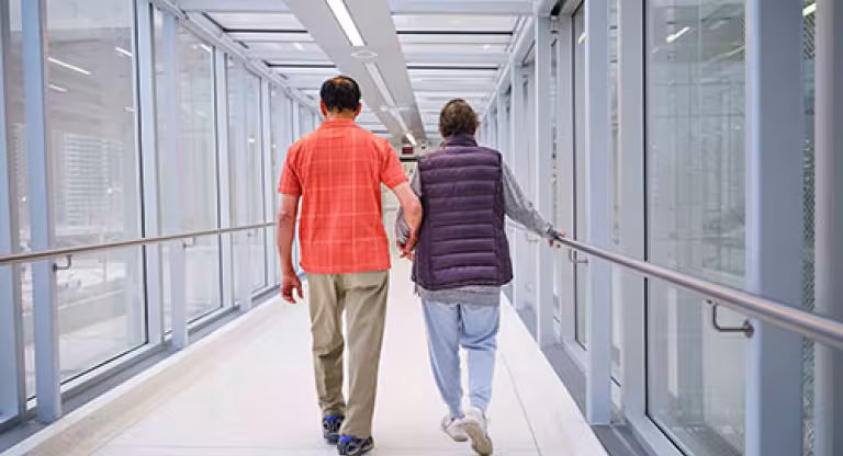 Medium long shot a man, wearing an orange checked shirt and khaki pants, holds hands with a woman, wearing a purple vest and grey sweat pants. They are walking down a hallway with windows with their backs to the camera. 
