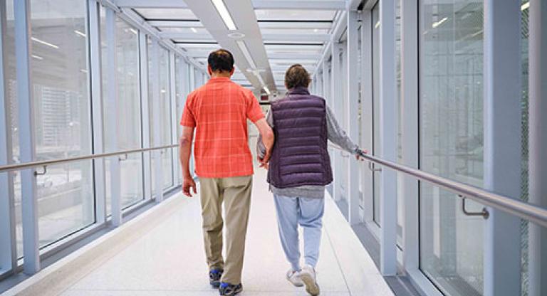 Medium long shot a man, wearing an orange checked shirt and khaki pants, holds hands with a woman, wearing a purple vest and grey sweat pants. They are walking down a hallway with windows with their backs to the camera. 