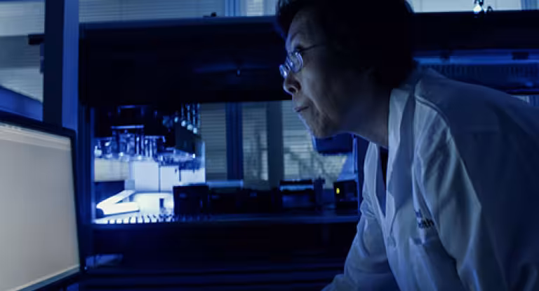 Medium close image of a woman wearing glasses and a white lab coat, standing in a dark room and leaning forward to read something on a glowing computer screen.