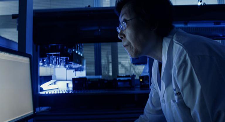 Medium close image of a woman wearing glasses and a white lab coat, standing in a dark room and leaning forward to read something on a glowing computer screen.