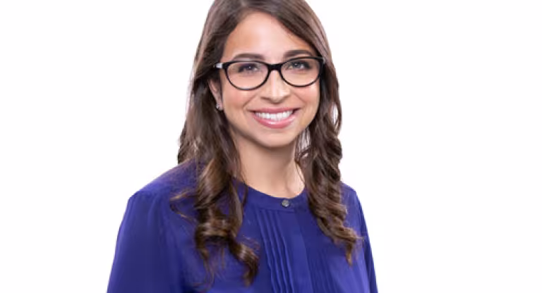 Medium close shot of a smiling woman with long, curled brown hair, wearing a purple blouse and black glasses