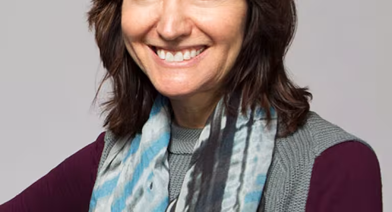 Medium close shot of a woman with shoulder length brown hair, wearing a blue and grey scarf, a knit grey vest and maroon shirt, is smiling.  