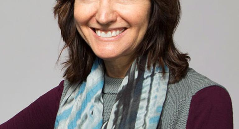 Medium close shot of a woman with shoulder length brown hair, wearing a blue and grey scarf, a knit grey vest and maroon shirt, is smiling.  