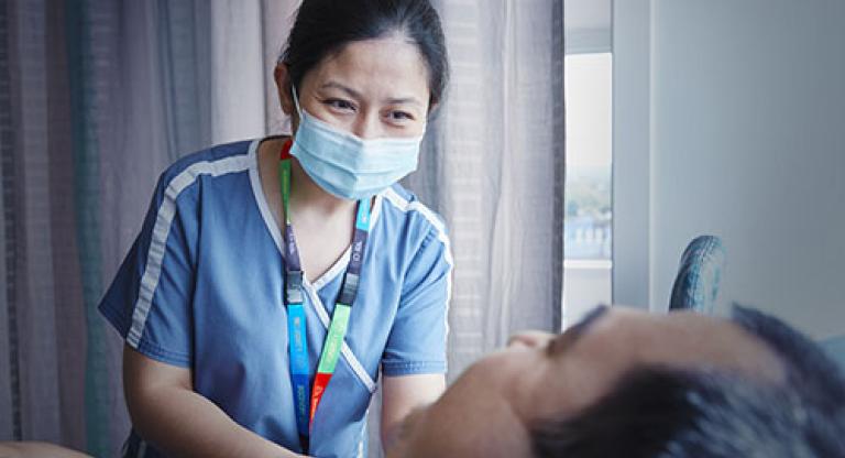 An Asian nurse wearing blue scrubs, a mask, and a colourful lanyard leans towards a man who is laying down. 