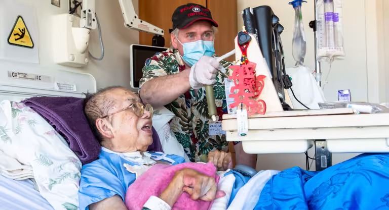 A woman in a hospital bed with her husband standing next to her. They are both looking at a computer screen and he is tapping the screen with a stylus