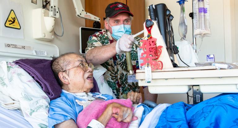 A woman in a hospital bed with her husband standing next to her. They are both looking at a computer screen and he is tapping the screen with a stylus