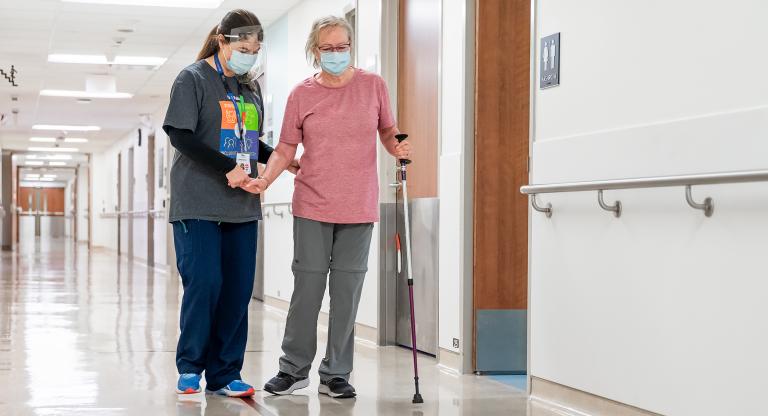 A health-care provider and a patient are walking down the hallway of a hospital. The patient is an older adult woman and she is walking towards the camera with a walking pole in her left hand. The health care provider walks on the patient's right side, holding her hand and offering support