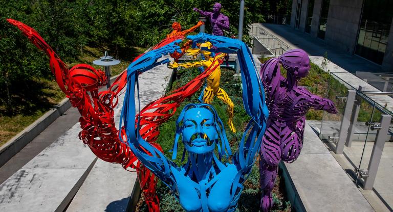 a group of six metal sculptures in the Max Tannebaum Sculpture garden. They are human figures made out of narrow strips of metal in bright colours. Each figure is depicted as if it is frozen in the middle of motion. The sculpture closest to the camera is dancing