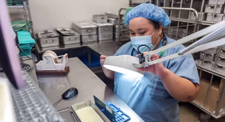 a woman wearing scrubs and a mask looks at a piece of surgical equipment through a magnifying glass