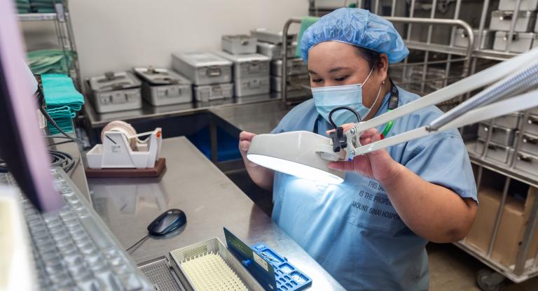 a woman wearing scrubs and a mask looks at a piece of surgical equipment through a magnifying glass