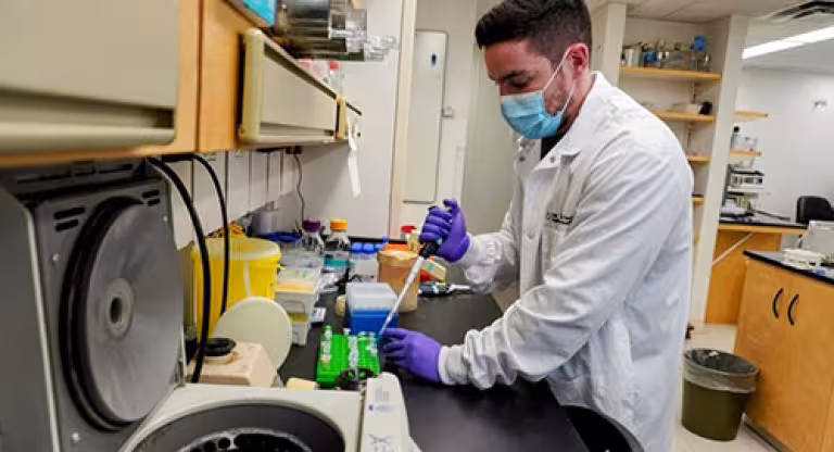 Medium close image of a man wearing a mask, a white lab coat, and purple gloves. He is transferring a substance with a pipette and there is a centrifuge in the foreground/