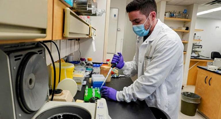 Medium close image of a man wearing a mask, a white lab coat, and purple gloves. He is transferring a substance with a pipette and there is a centrifuge in the foreground/