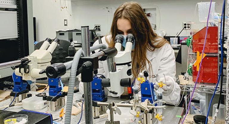 Medium close image of young woman with long hair sitting at a medical work station surrounded by equipment, looking into a microscope.