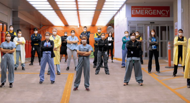 A large group of health-care workers standing outside of the Emergency department with their arms crossed. 