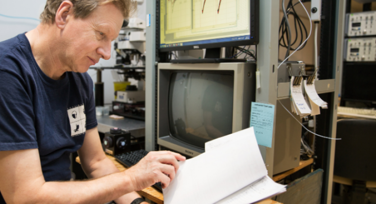 Graham Collingridge reads a printed document while sitting in front of a computer monitor.
