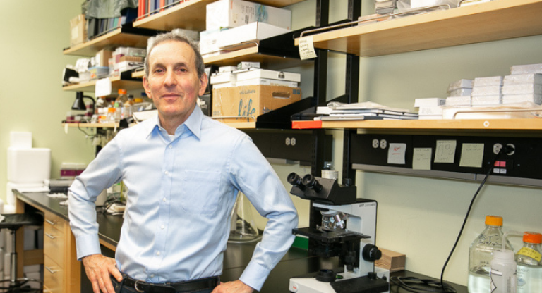 Dr. Daniel Drucker in a blue dress shirt standing in a lab. 