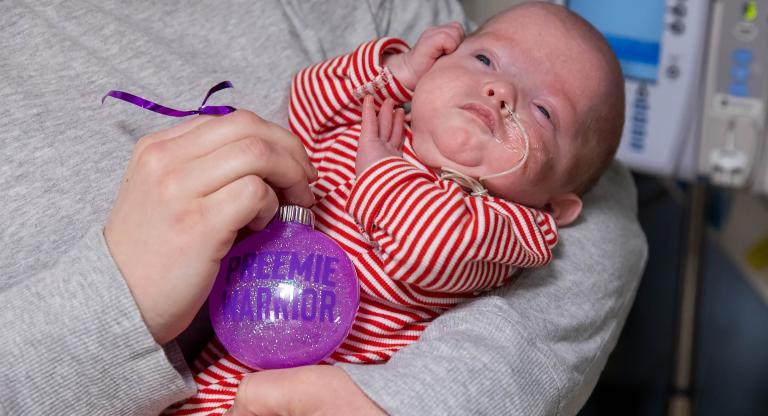 Infant in the NICU held in their mother’s arms beside a hand-made ornament. Caption: This holiday season, families in our NICU received hand-made, donated ornaments.