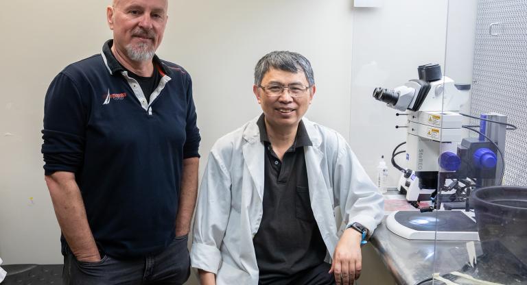 Two researchers pictured in the laboratory next to a microscope. One researcher. A Caucasian man is standing with hands in pockets and the other, a bespectacled Asian man, is seated and wears a lab coat.