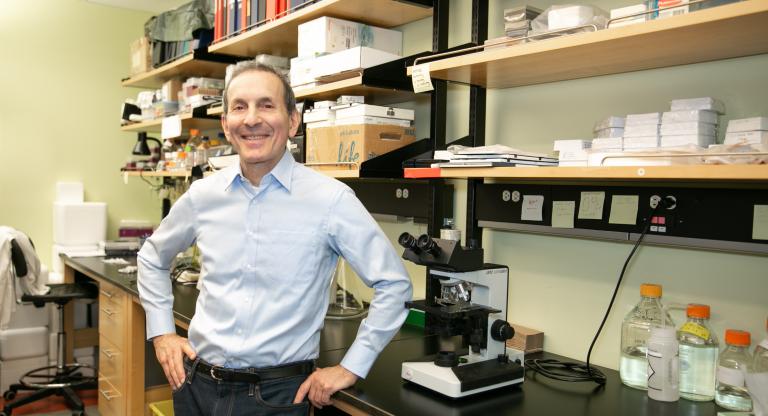Dr. Daniel Drucker in a light blue dress shirt standing in his lab with a smile.