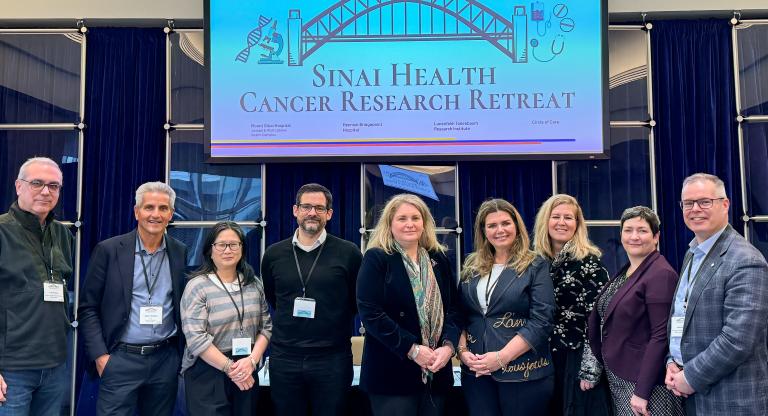 A group of nine health-care professionals standing together in a single row, facing the camera smiling. They are wearing business attire. They are standing under a screen with an image of a bridge and the words, Sinai Health Cancer Research Retreat