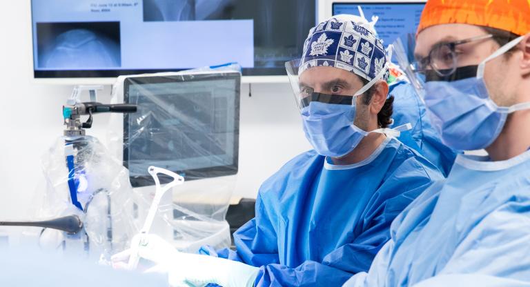 Dr. Jesse Wolfstadt in a blue gown and mask using advanced robotic equipment in a sterile operating room.