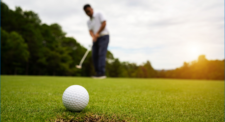 Close-up of a golf ball near the hole with a golfer putting in the background.