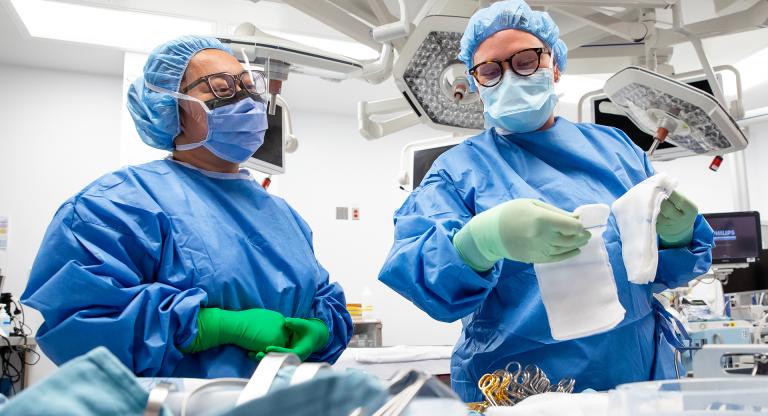 Two nurses stand in front of a table of surgical instruments in an Operating room. The nurses are wearing scrubs, including blue gown, gloves, caps and face shields. One nurse is holding out surgical sponges while the other watches