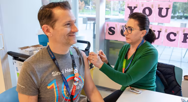 A woman prepares to give a vaccination to a man. They are both sitting at a table that is in front of a window.
