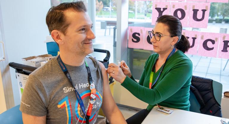 A woman prepares to give a vaccination to a man. They are both sitting at a table that is in front of a window.