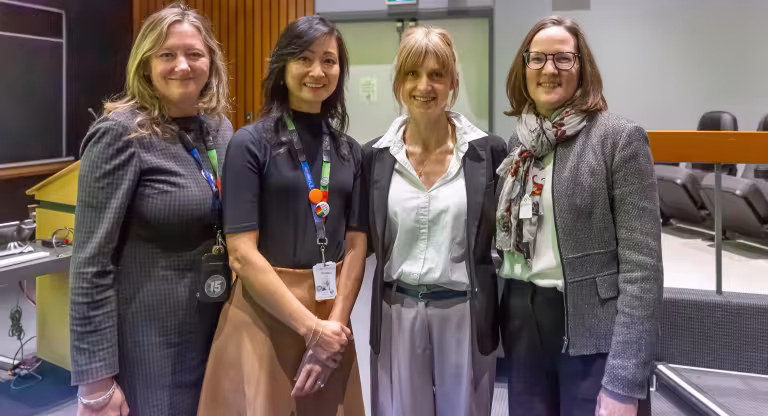 A group of four women standing in a line, side-by-side. They are looking at the camera, smiling. They are in an auditorium