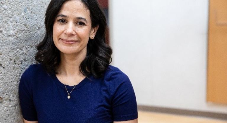 Dr. Amanda Selk stands indoors beside a textured concrete pillar, smiling softly at the camera while wearing a navy blue top and small gold necklace. The background shows a bright hallway with light flooring and neutral walls.