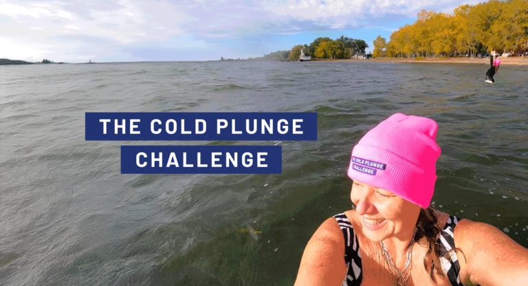 A person wearing a bright pink winter hat stands in cold lake water near a shoreline lined with autumn trees. Another participant is visible farther out on the right. Text over the water reads “The Cold Plunge Challenge” against a blue background.