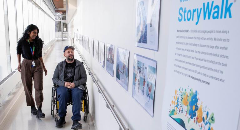 A patient using a wheelchair moves along a hospital hallway with an occupational therapist, viewing the Story Walk panels displaying illustrated book pages mounted on the wall at Hennick Bridgepoint Hospital.