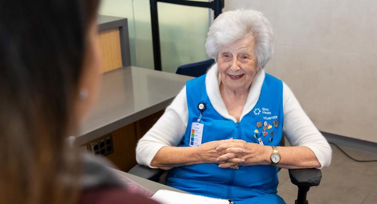A woman sits at an information desk at a hospital greeting a patient with a smile on her face. 