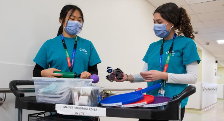 Two Sinai Health co-op students wearing teal scrubs and masks stand beside a cart stocked with recreational items.