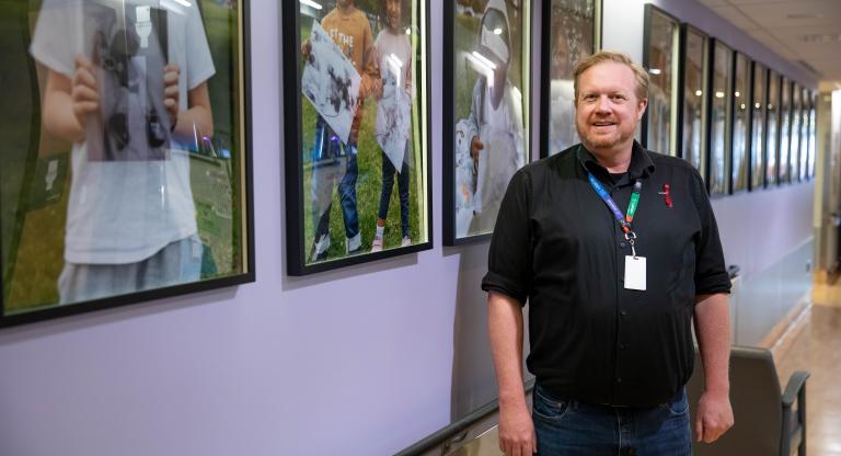 Patient Care Manager, John Balcom, stands and smiles in a light purple hospital hallway beside a series of framed photos on the NICU Hero Wall at Mount Sinai Hospital.
