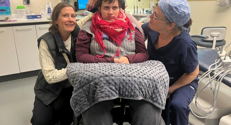 Young woman sits in a wheelchair in a dental clinic, with her dentist and mother smiling beside her
