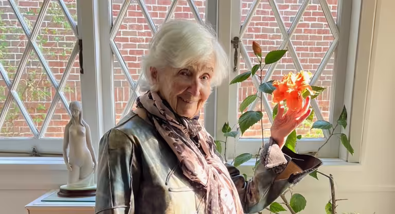 elderly woman stands by a window holding a flower