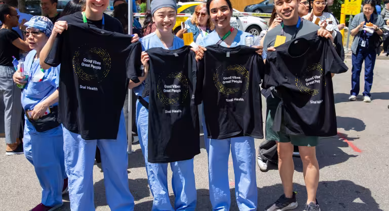 Four Sinai Health employees stand outdoors at the People Appreciation BBQ, smiling and holding black “Good Vibes Great People” T-shirts.