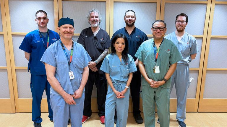 Group of people wearing scrubs and colourful lanyards stand in front of a wall smiling.