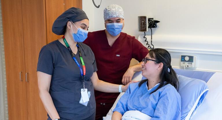 Patient smiles while seated in a hospital bed, interacting with two masked health professionals
